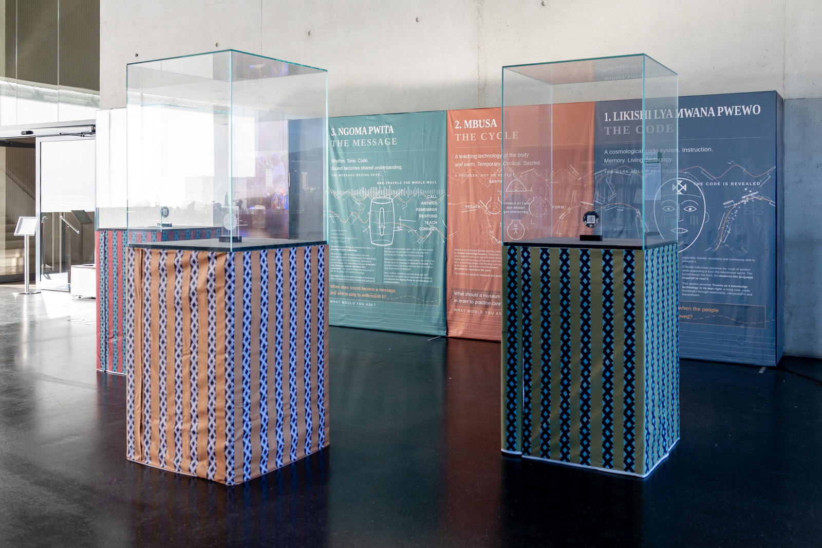 Three empty glass vitrines on fabric-wrapped plinths stand in front of the exhibition wall: blue, terracotta and teal banners for Likishi Lya Mwana Pwewo, Mbusa and Ngoma Pwita.
